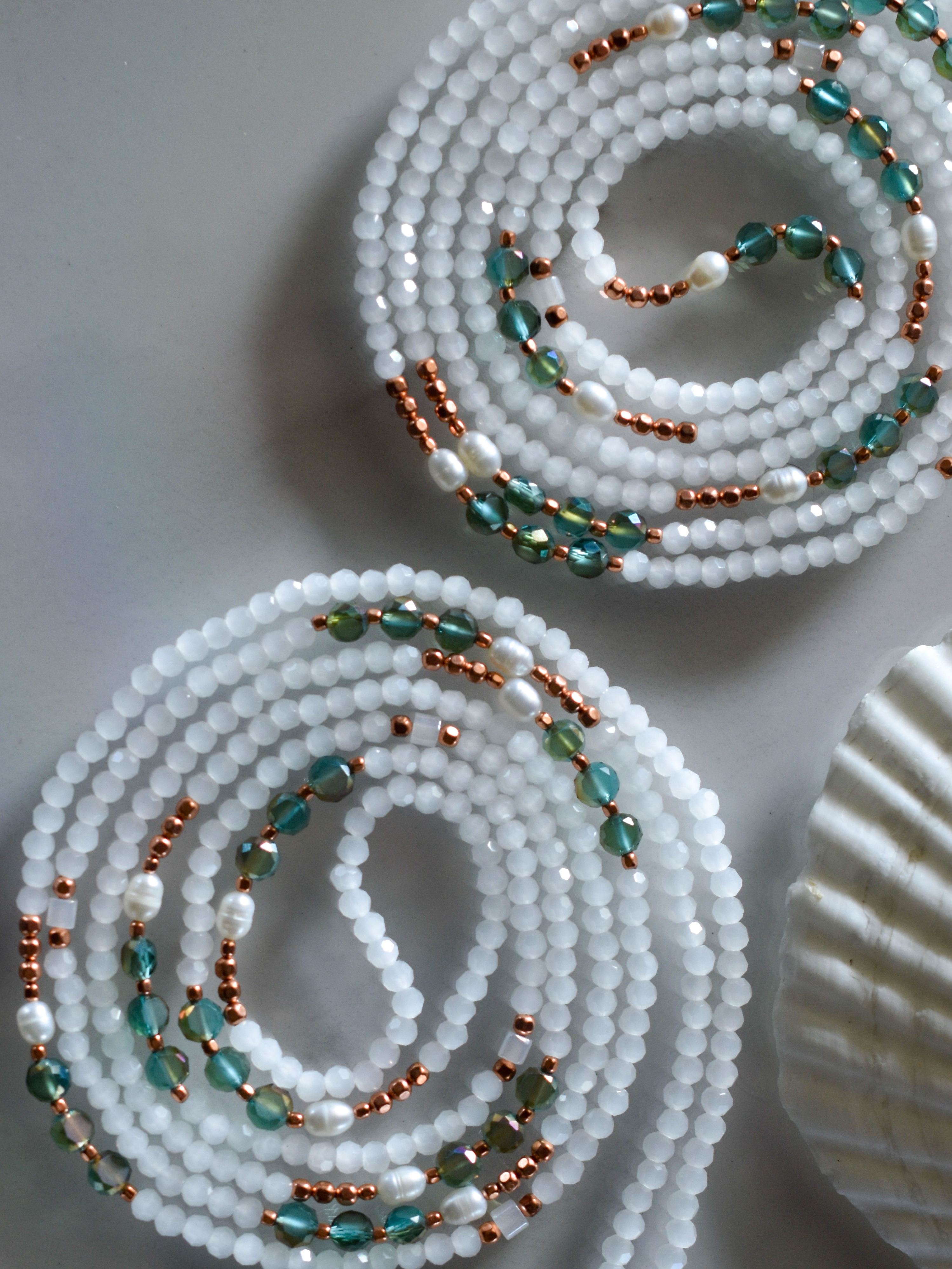 Two spirals of waist beads featuring white faceted beads, teal/green crystal beads, small copper seed beads, and white irregular pearls.