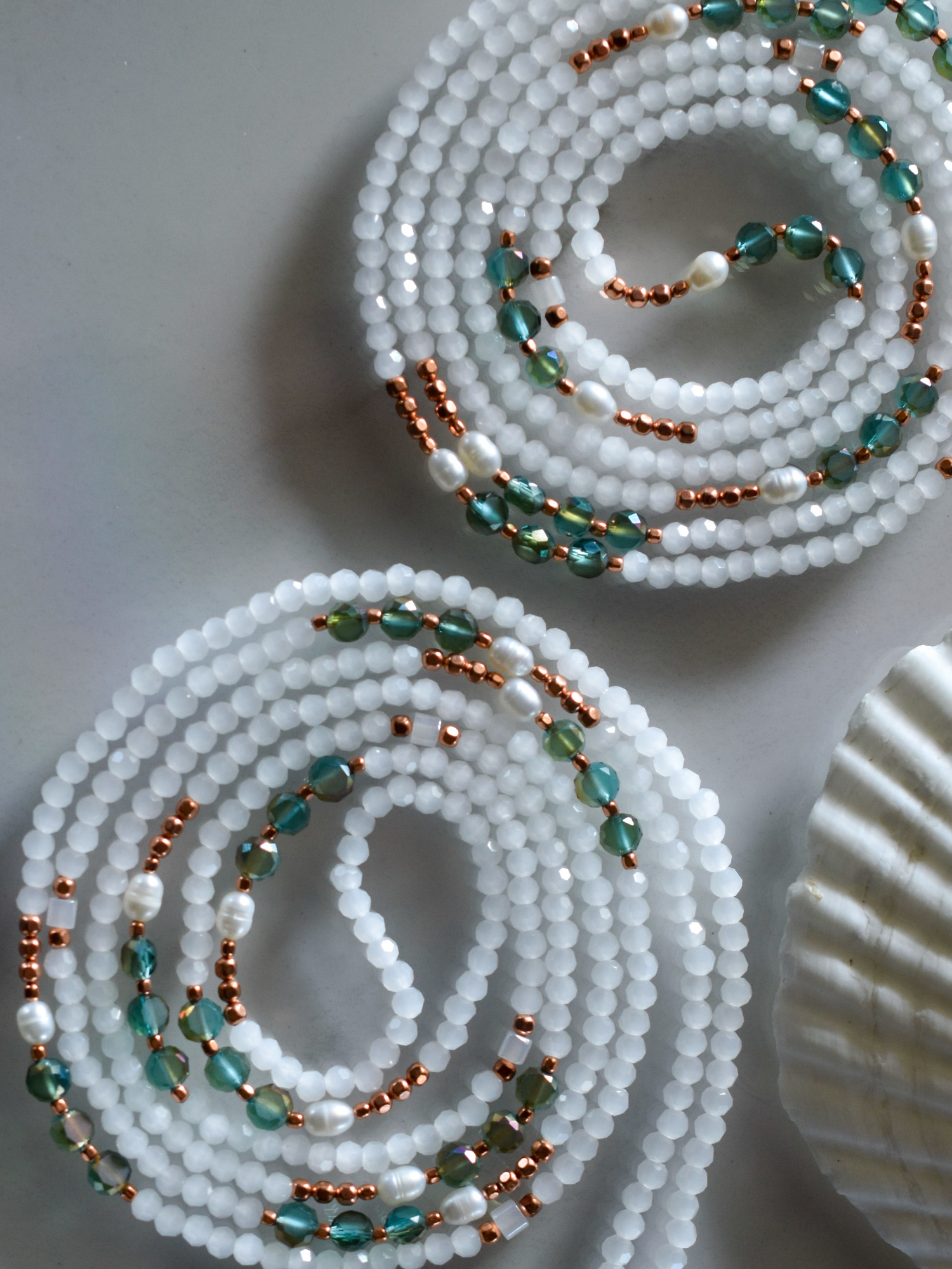 Two spirals of waist beads featuring white faceted beads, teal/green crystal beads, small copper seed beads, and white irregular pearls.